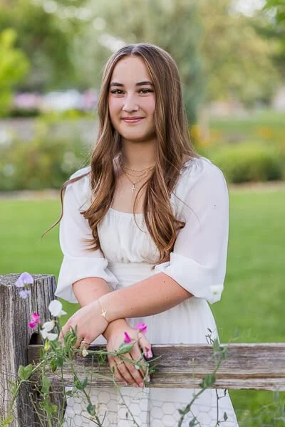 Teen girl in white dress leans against flower-lined picket fence