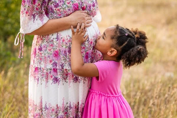 Little girl in pink dress kisses mom's baby bump
