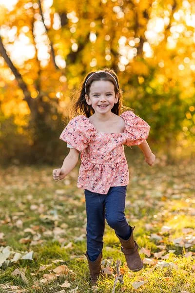 Little girl in pink printed shirt & jeans runs joyfully through fall leaves
