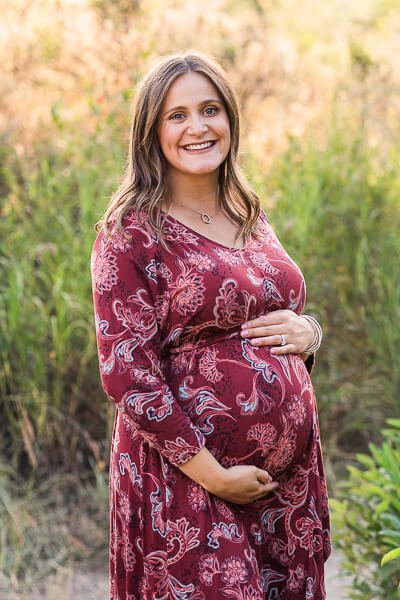 Pregnant woman in paisley burgundy dress smiles as she cradles her baby bump