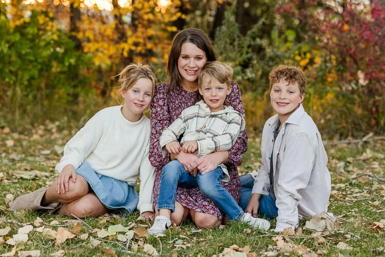 Mom & 3 kids sit on the ground together surrounded by fall foliage
