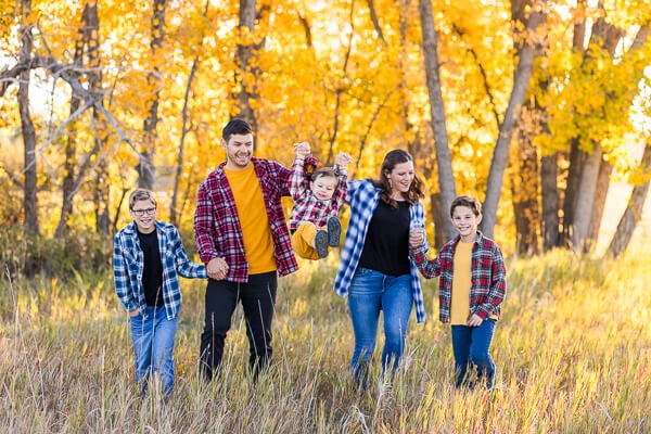 Family of 5 walks through fall foliage holding hands, with parents swinging toddler in between them