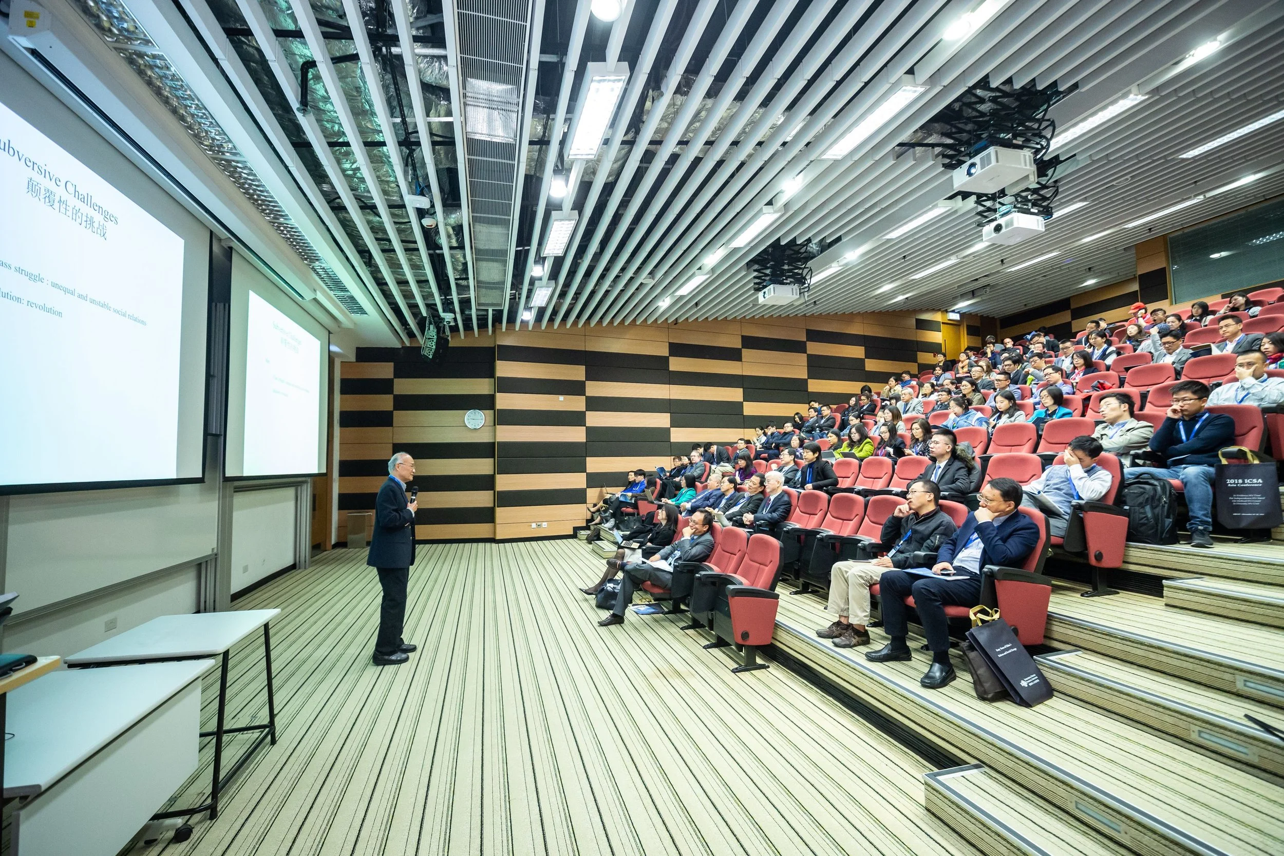 A man speaking at a conference, standing in front of a large audience seated in red chairs. The auditorium has a modern design with striped wood panel walls, ceiling-mounted projectors, and a large presentation screen displaying text.