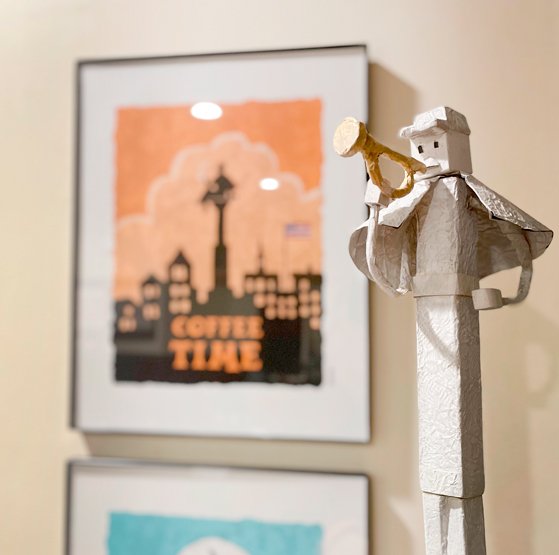Close-up of a paper sculpture of a musician playing a trumpet, with a blurred background featuring framed artwork of a skyline and the words "Coffee Time."