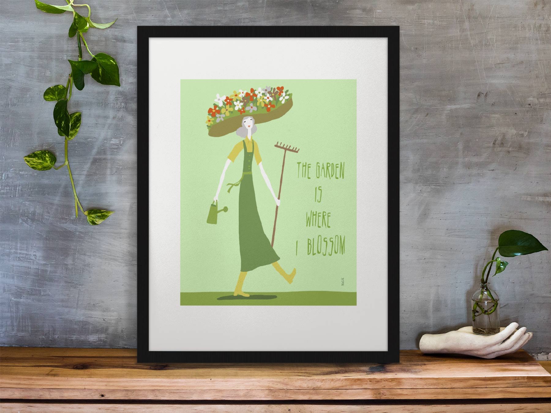 framed-art-print” I-Am-The Garden”-standing-on-a-wooden-desk-against-a-concrete-wall-near-a-hand-decoration.jpg