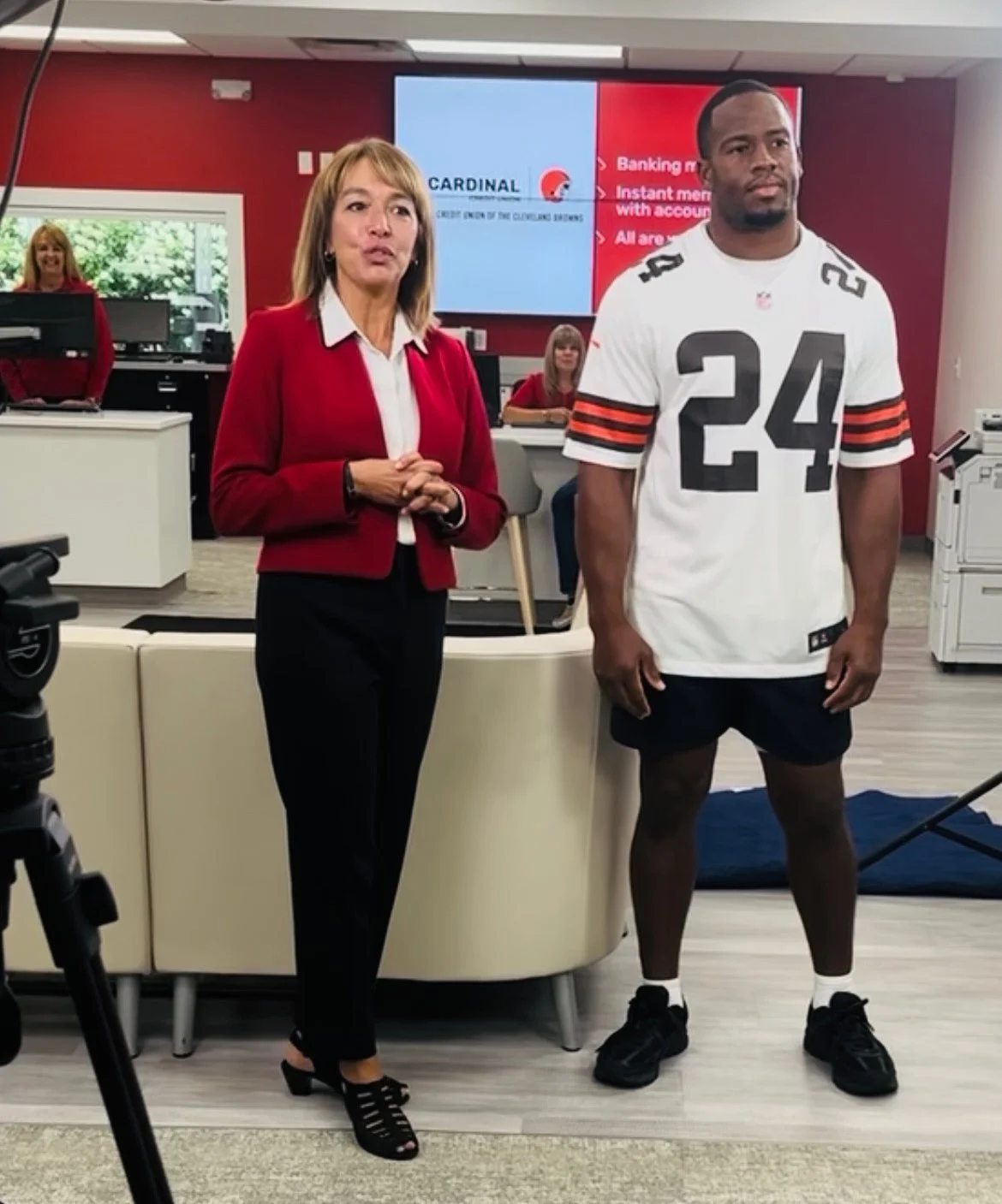 A woman in a red blazer and black pants standing next to a man in a Cleveland Browns football jersey and shorts inside a bank or office setting.