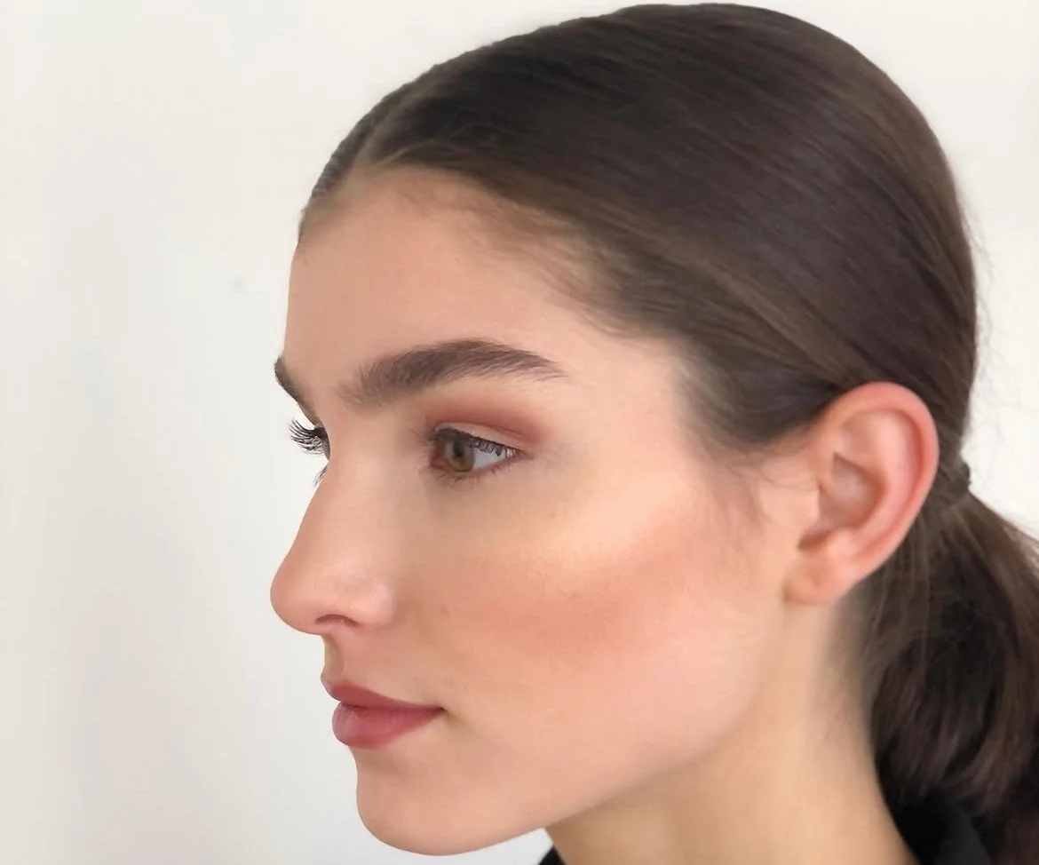 Close-up profile of a woman with brown hair pulled back, wearing natural makeup, against a plain white background.