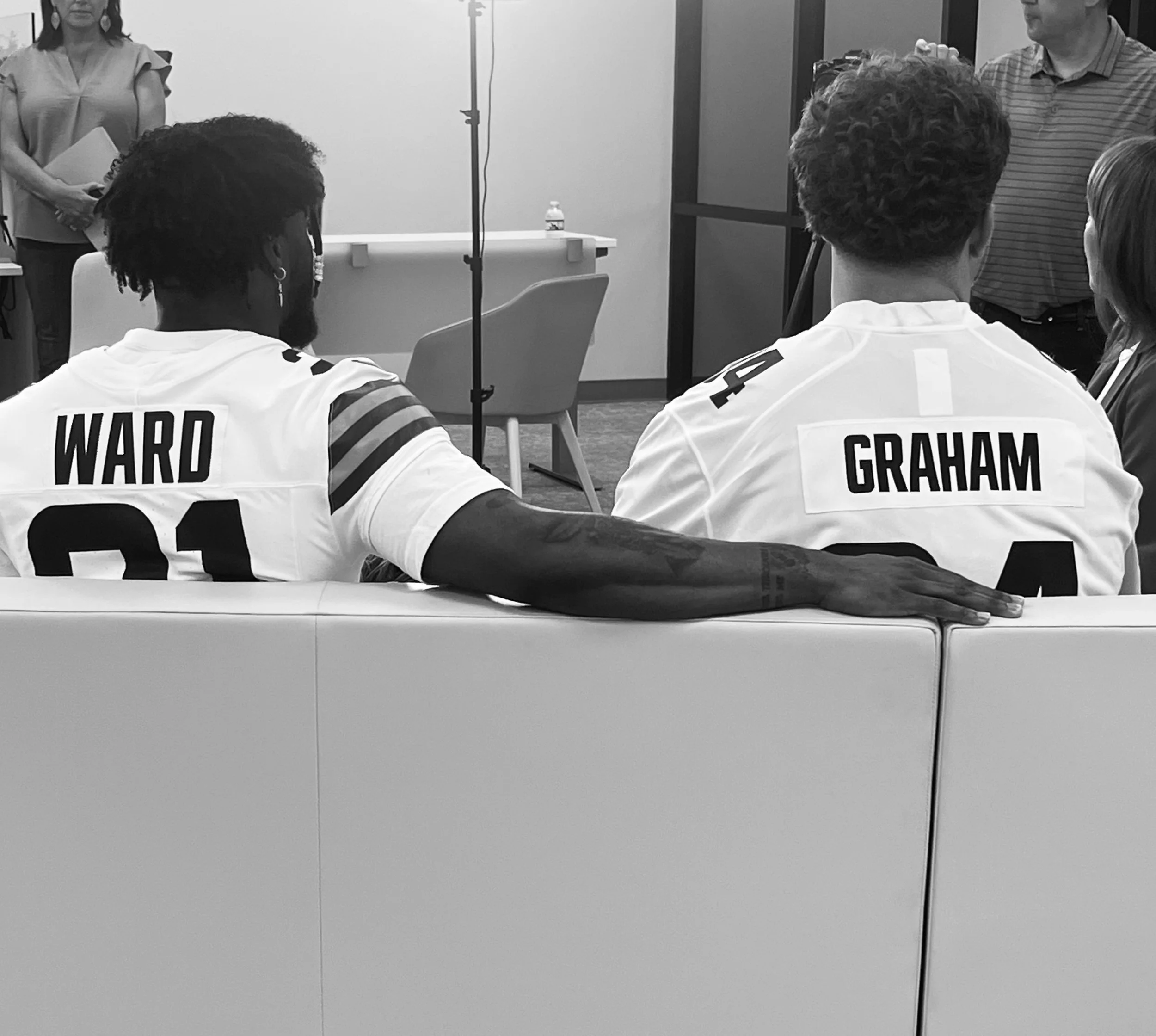 Two football players wearing jerseys with the names Ward and Graham sitting on a sofa, with a woman standing and holding a folder and two individuals standing in front of them in a room.