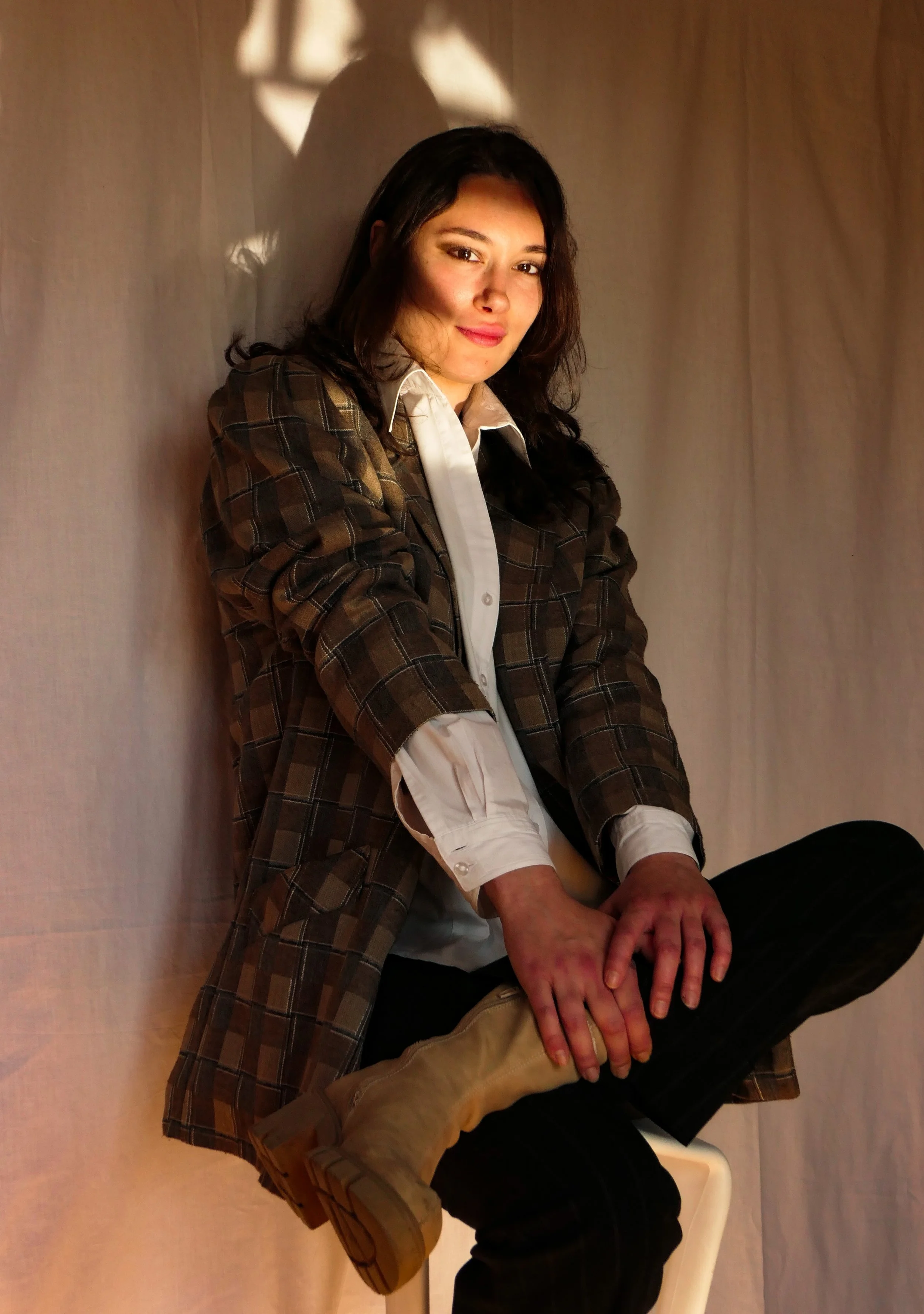 A woman with dark brown hair, sitting against a beige background, wearing a plaid blazer, white shirt, black pants, and beige boots, posing with a relaxed smile.