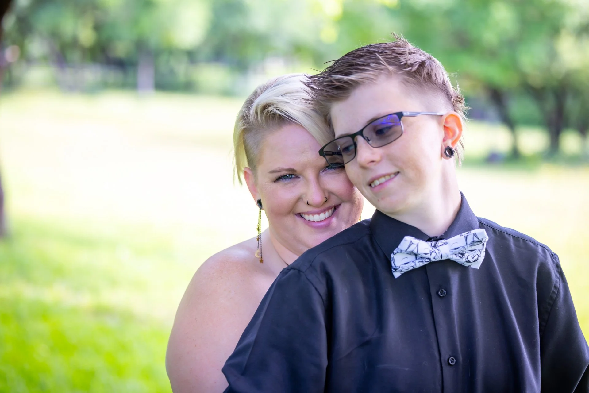 A woman with short, platinum blonde hair and a vertically shaved section on the side, smiling and leaning her head against a young person with short brown hair, glasses, and pierced ears, near a park with trees and greenery in the background.