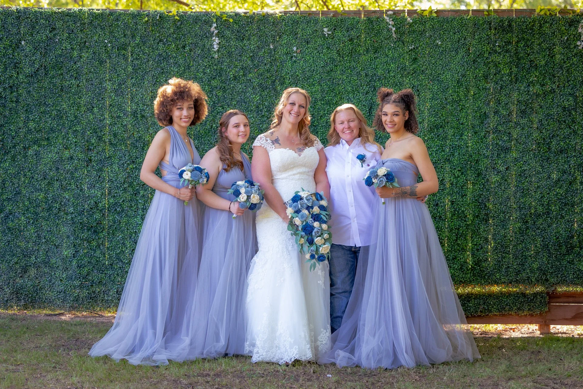 Group of five women standing outdoors in front of a green hedge, with the bride in a white wedding dress and four bridesmaids in matching lavender dresses, all holding bouquets of white and blue flowers.