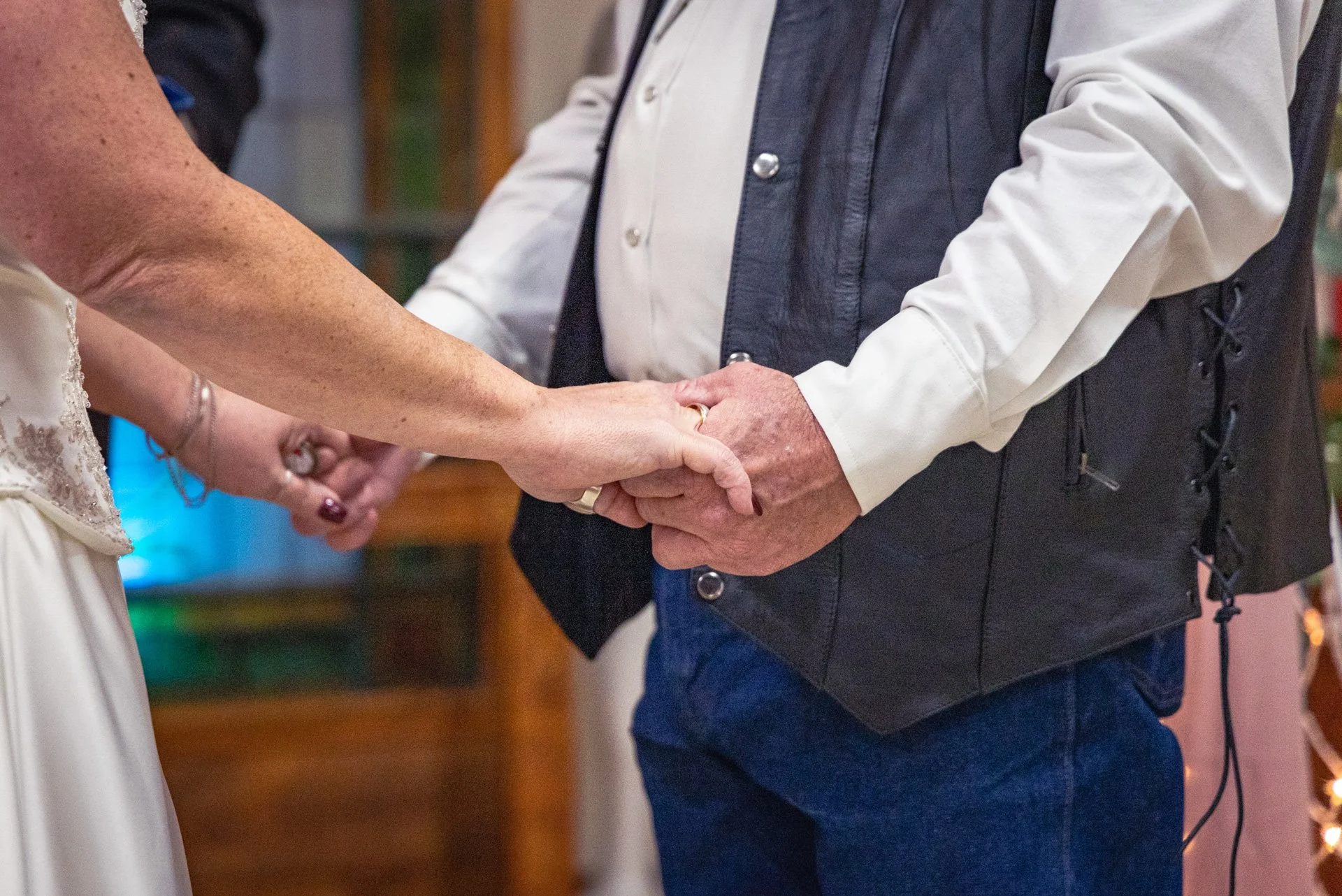 A couple holding hands during a wedding ceremony, with the bride wearing a white dress and the groom in a white shirt and dark vest, indoors with warm lighting.