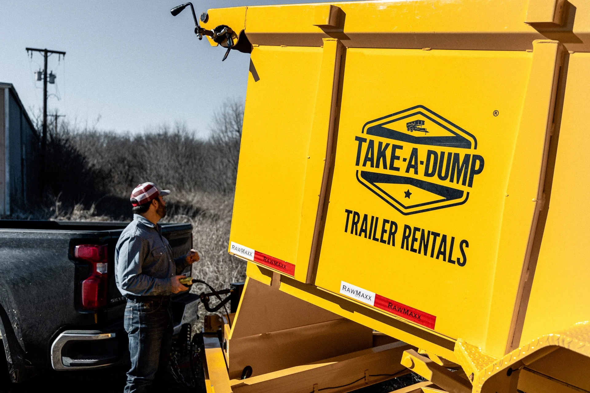 A man in a plaid cap and gray jacket standing beside a bright yellow trailer with the words "Take-A-Dump Trailer Rentals" written on it, attached to a pickup truck on a rural road.
