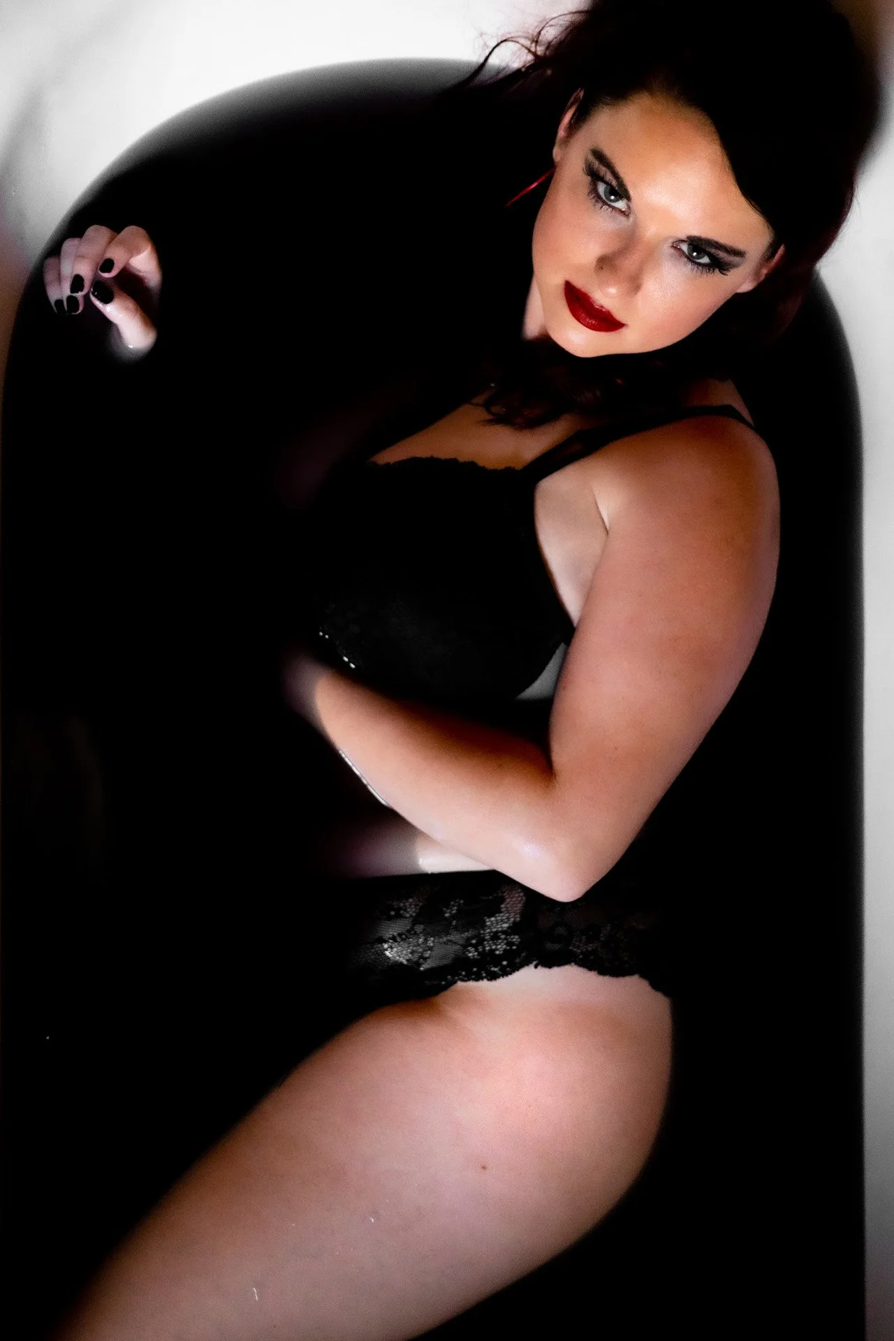 A woman with dark hair wearing black lace lingerie, sitting in a bathtub with a relaxed pose, looking up at the camera.
