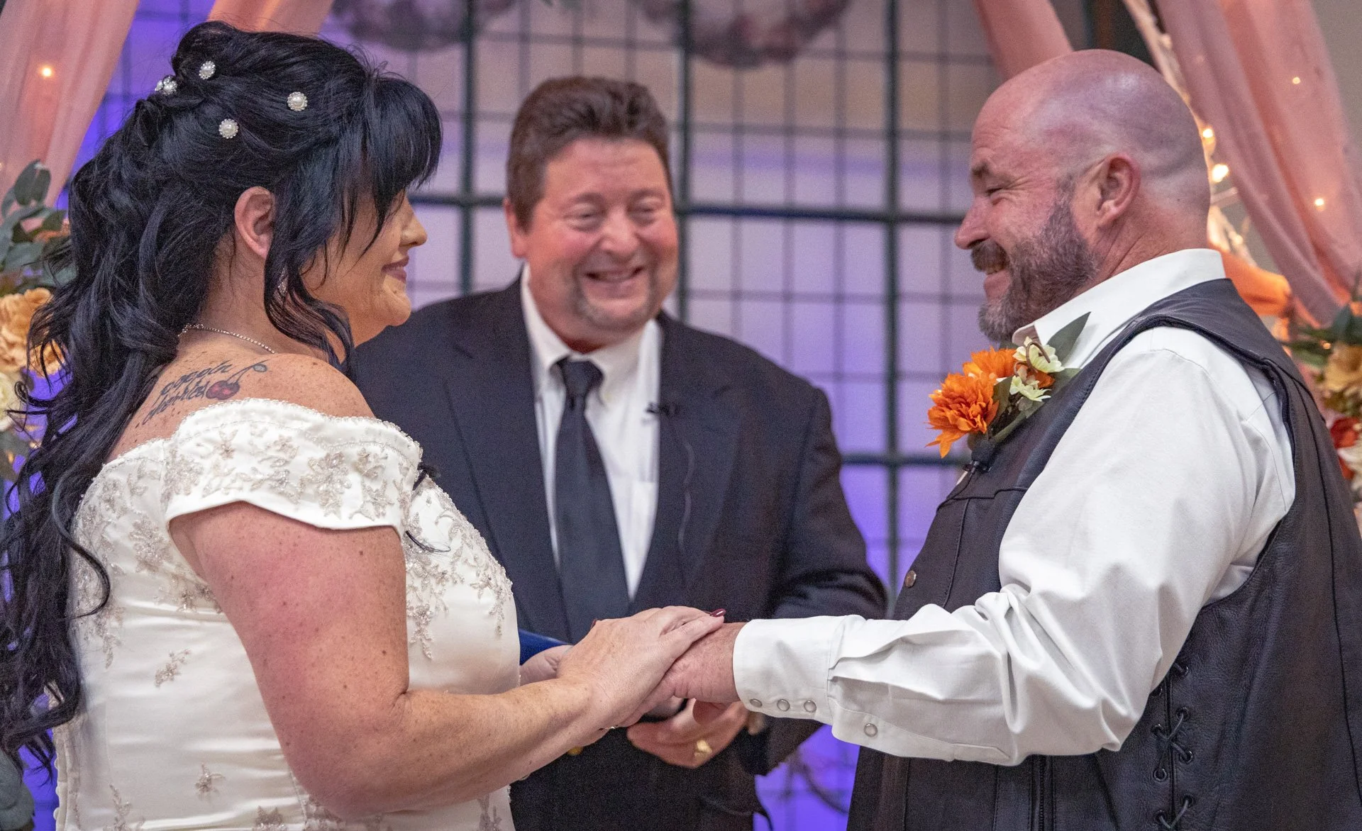 A same-sex wedding ceremony with two brides holding hands and smiling at each other, an officiant standing behind them smiling, and floral decorations in the background.