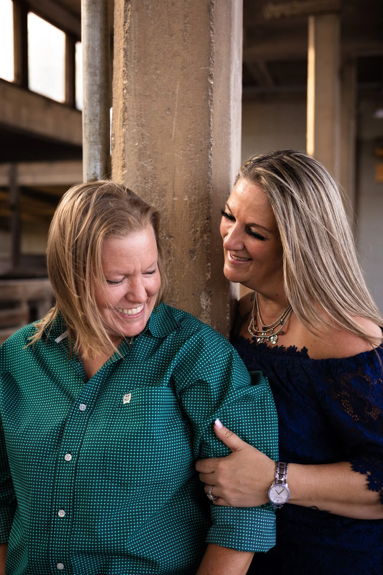 Two women are smiling and laughing, standing close together and leaning against a concrete pillar in an industrial-style setting.