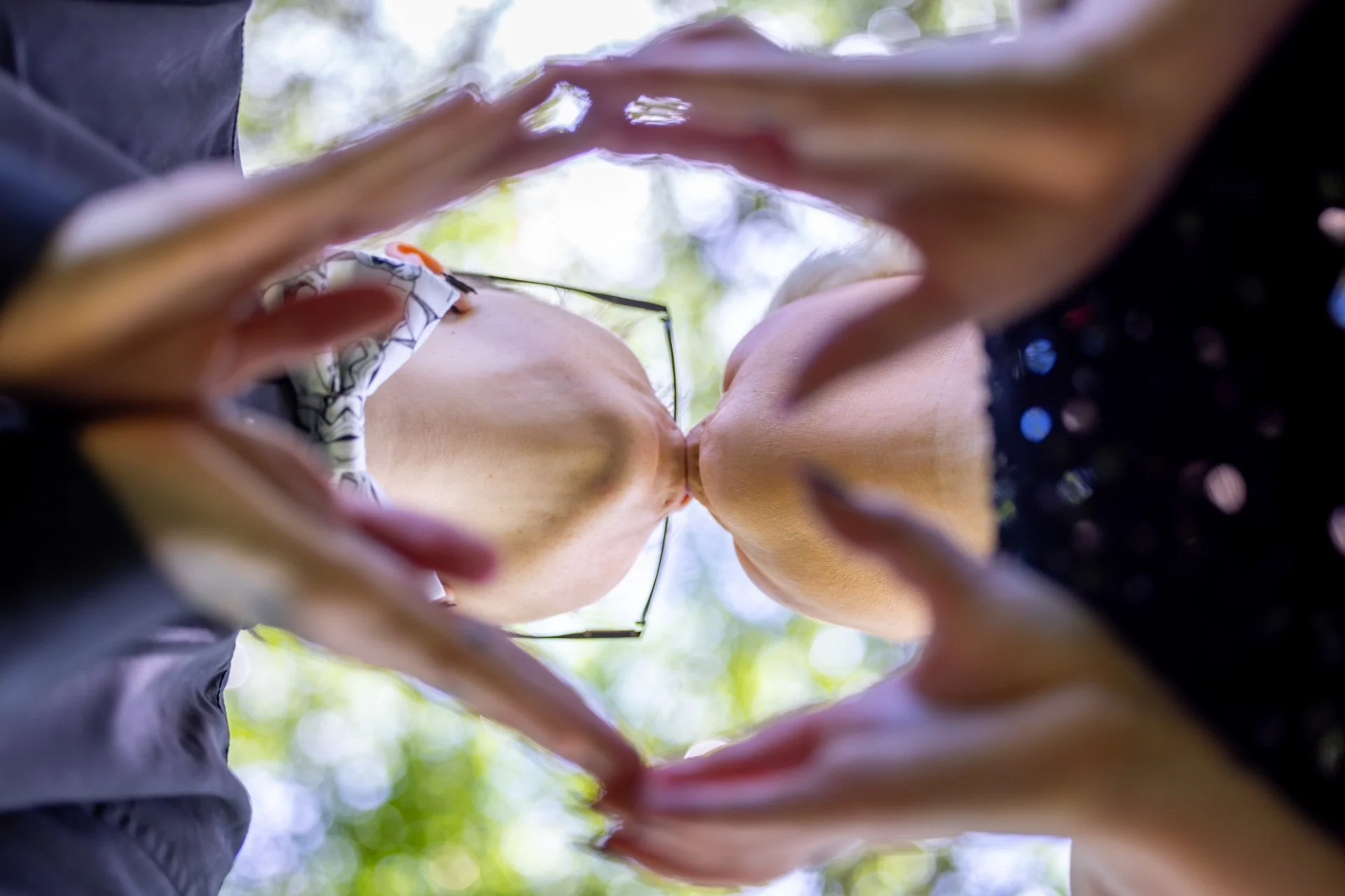 Low-angle close-up of two women kissing, forming a heart shape with their hands, outdoors with trees and sunlight in the background.