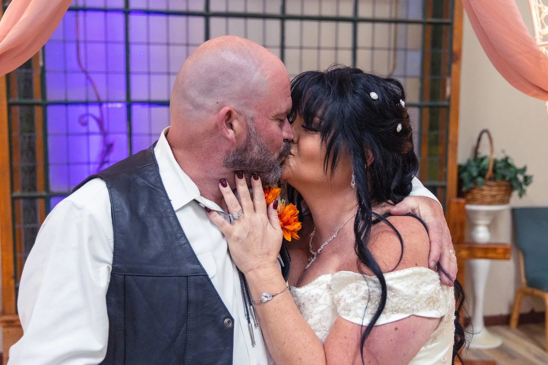 A man and woman kissing at a wedding or celebration, with the woman wearing a white dress and jewelry, and the man in a white shirt and leather vest.