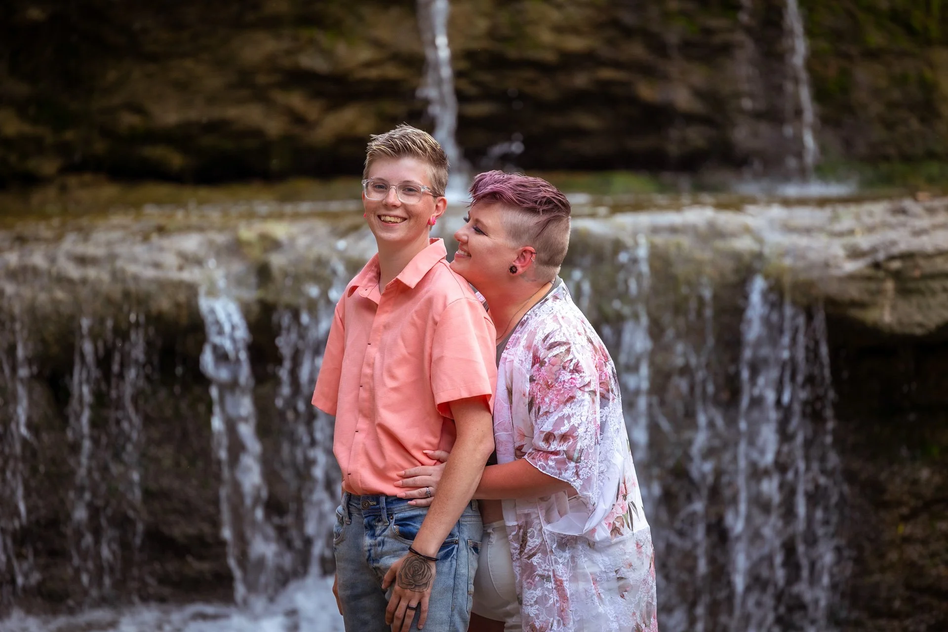Two women smiling and hugging near a small waterfall with mossy rocks in the background.