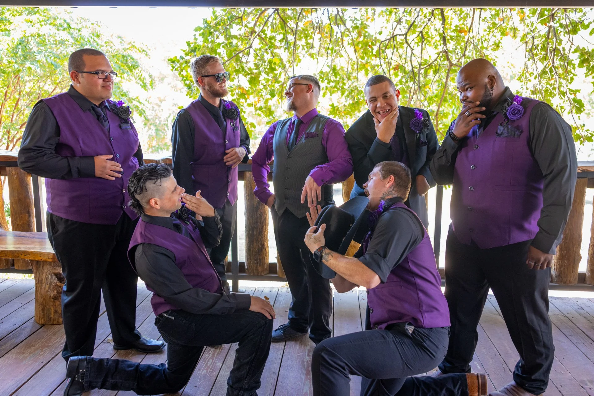 A group of men in matching purple and black formal attire gathered on a wooden deck with green trees in the background, engaged in playful conversation and laughter.