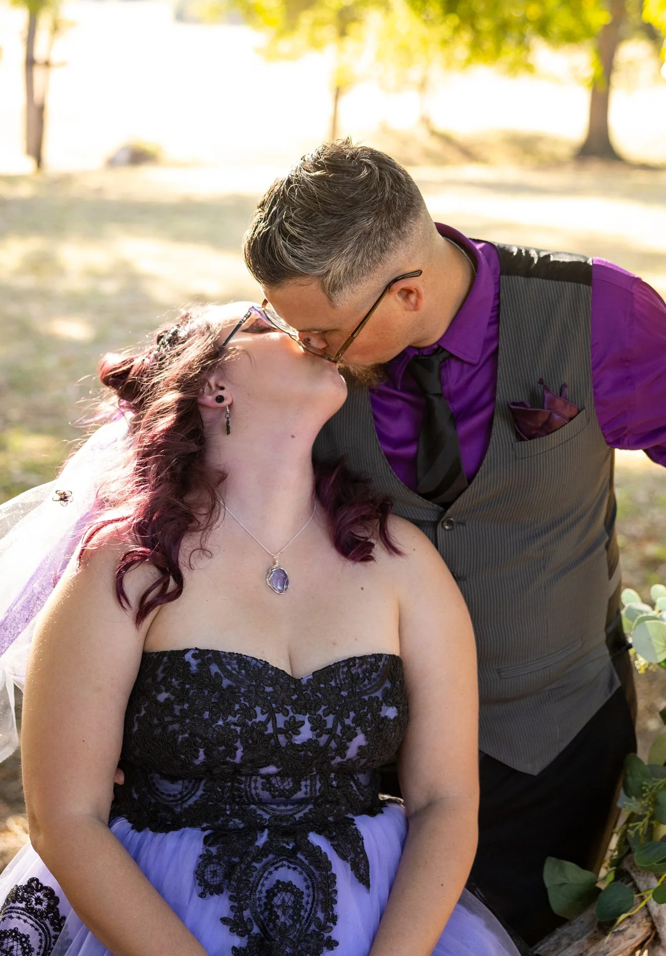 A couple sharing a kiss outdoors on a sunny day, with the woman wearing a strapless purple and black lace dress and a necklace, and the man wearing glasses, a purple shirt, a black vest, and a tie.