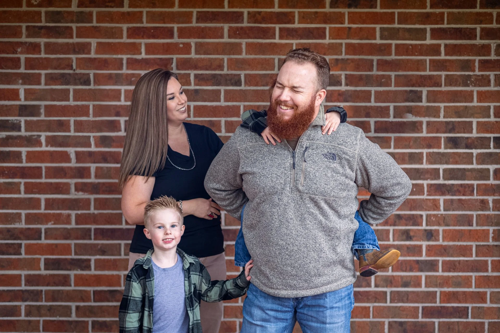 A family of four smiling against a brick wall. The father, with a red beard, is giving a piggyback ride to his young son, and the mother is standing beside them, smiling. The son is wearing a plaid shirt, and the daughter, who has her arms around her father's shoulders, is not fully visible but is on his back.