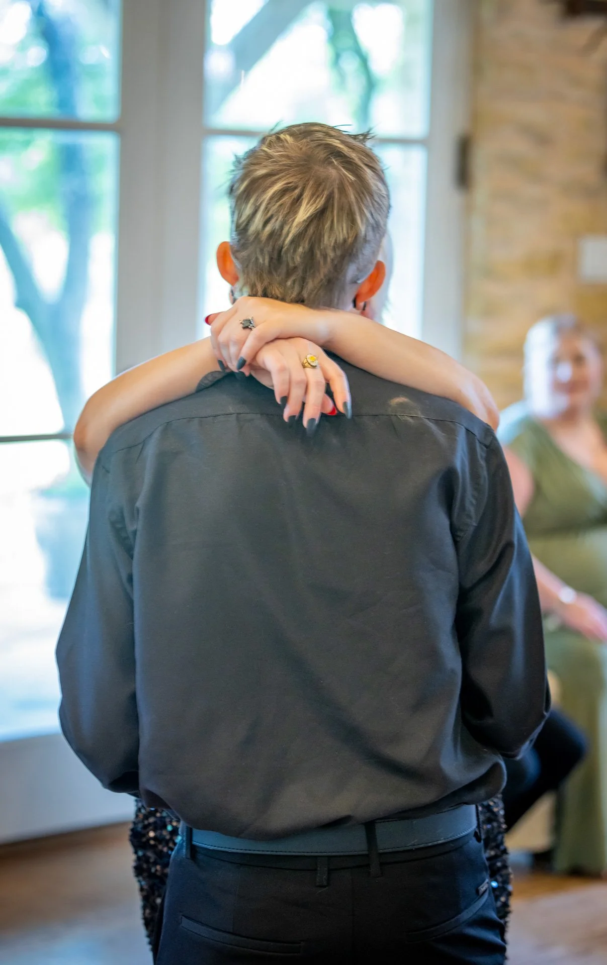 A woman with her arms around a man, hugging him from behind. The man is wearing a black shirt and is facing away from the camera, while the woman is looking at him, with part of her face visible in the background.