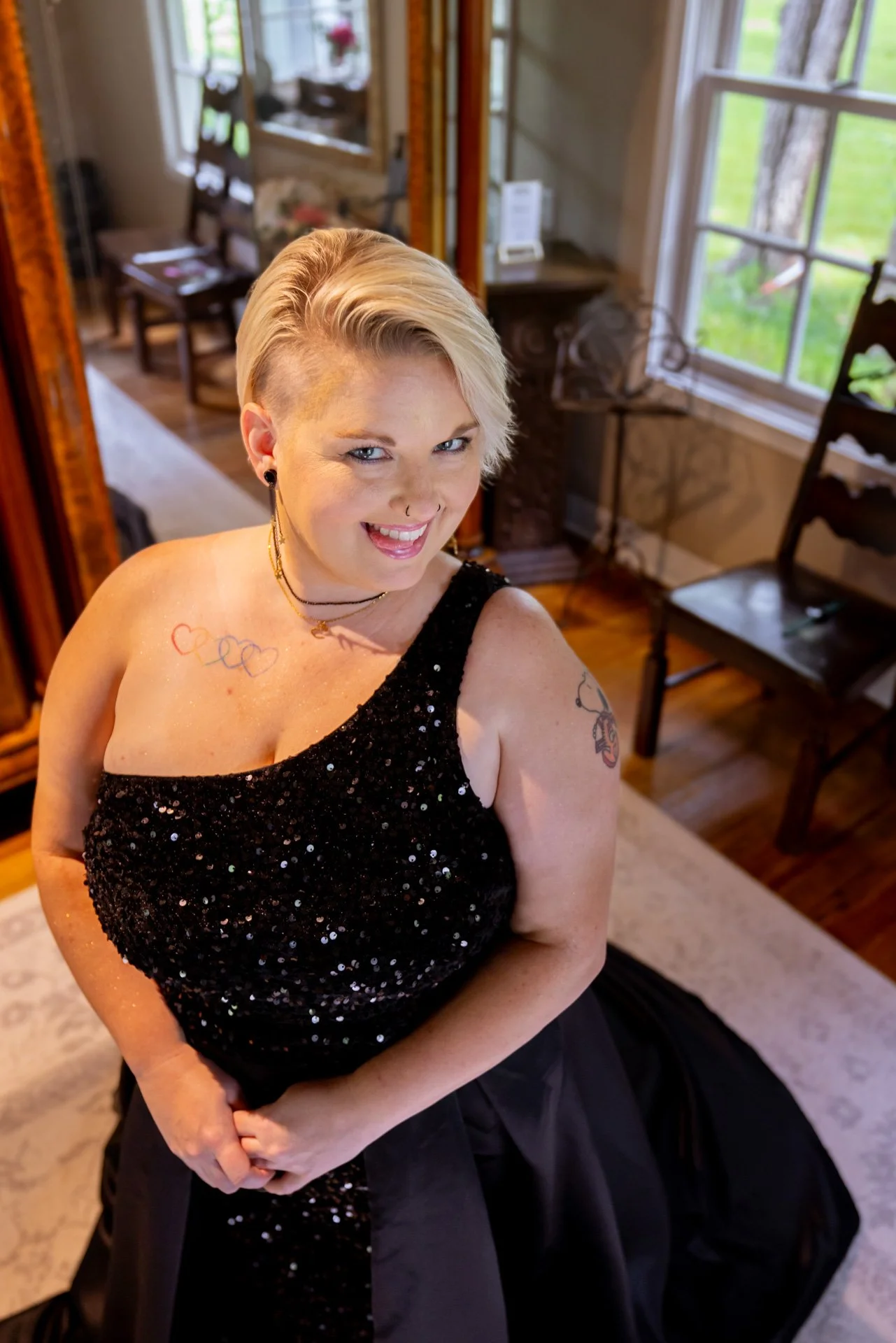 A woman with short blond hair, wearing a black sequin dress, smiling and standing in a room with wooden floors, chairs, and large windows showing green trees outside.