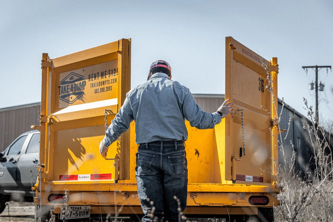 Man standing behind the open doors of a yellow dumpster truck, wearing a gray shirt and black jeans, on a sunny day with a gray car, power lines, and a warehouse in the background.