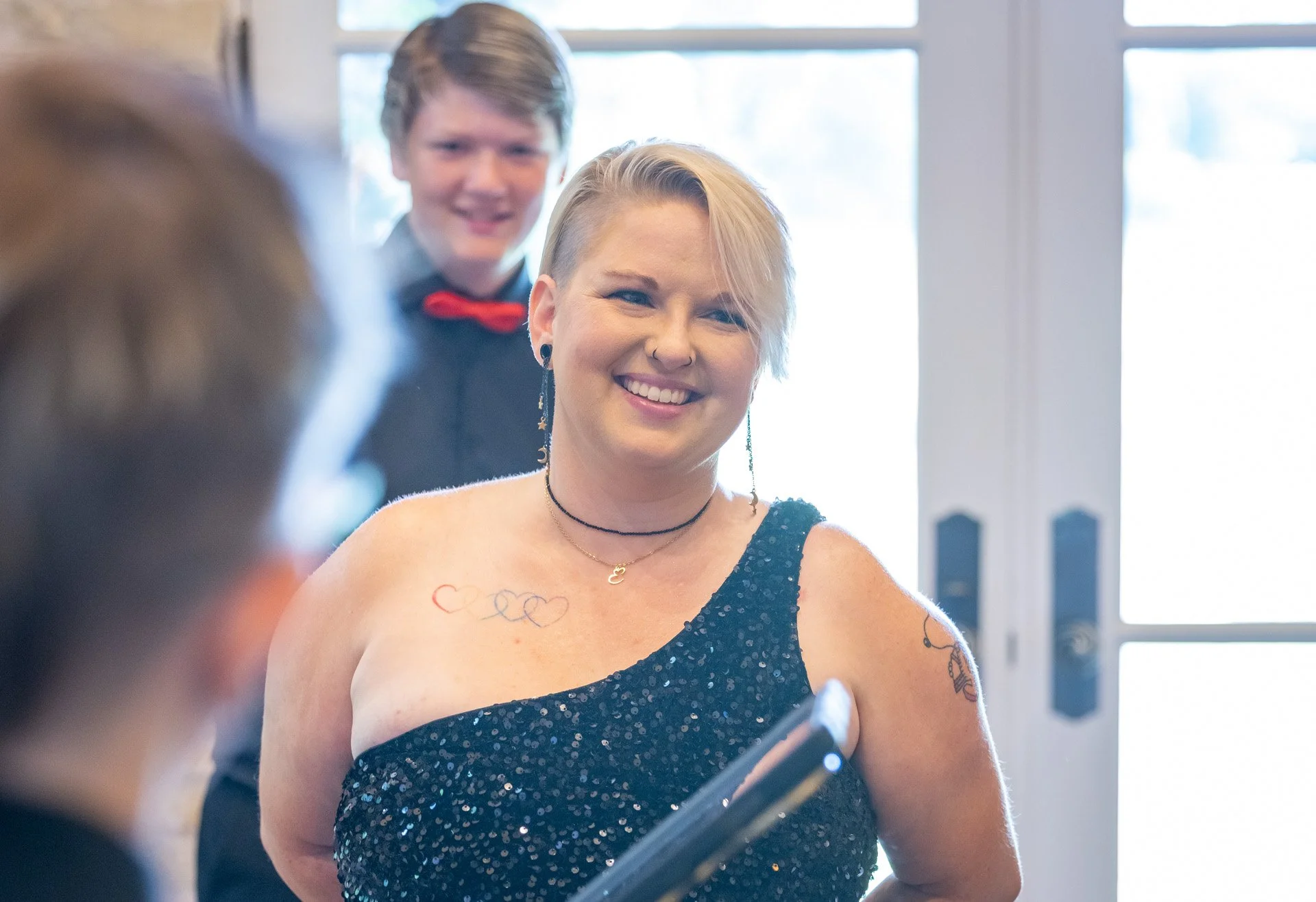 Woman with short blonde hair and tattoos smiling, wearing a black sequin dress, in a well-lit room with a man in the background and a person blurred in the foreground.