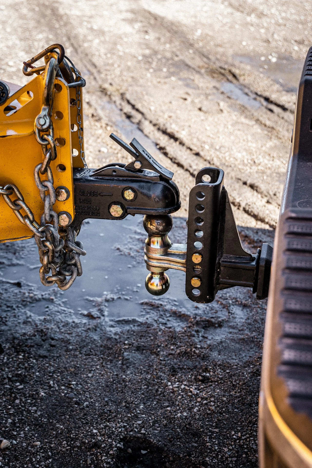 Close-up of a trailer hitch mechanism attached to a vehicle, with a black and silver hitch ball, metal chains, and a yellow safety chain, on a dirt surface.