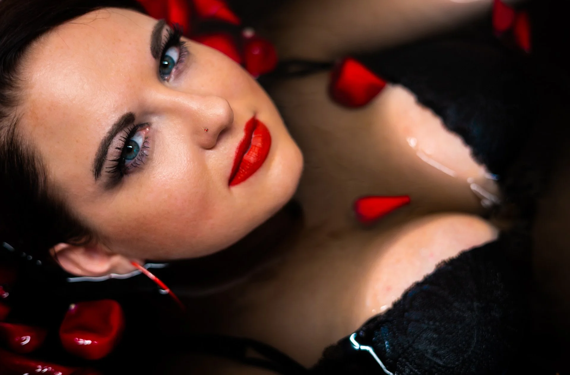 A close-up of a woman with blue eyes and red lipstick, lying down with red rose petals around her, wearing black lace clothing.