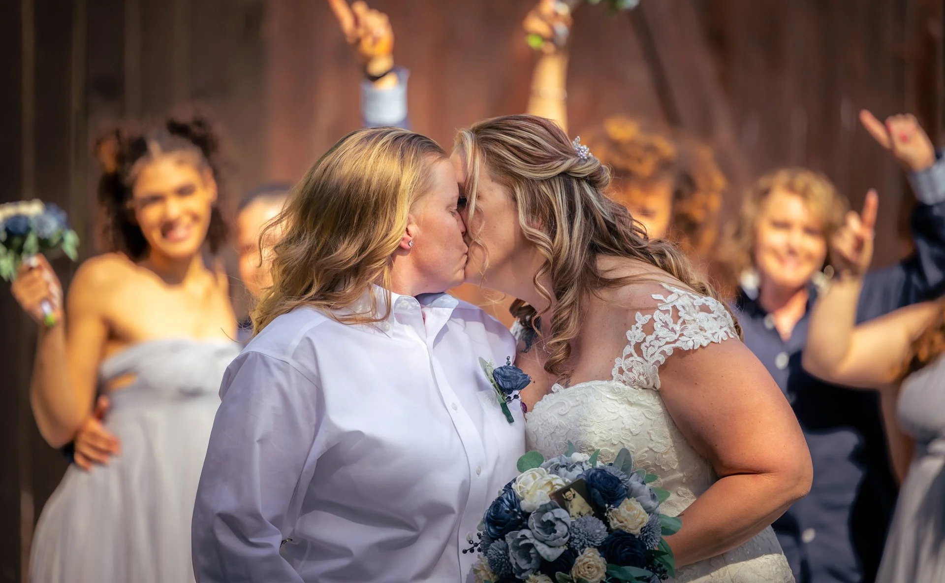 Two women in wedding attire sharing a kiss during a wedding ceremony, surrounded by smiling guests holding bouquets and raising their hands in celebration.