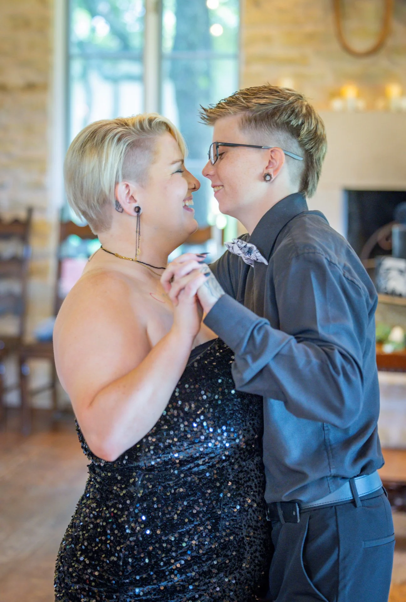 Two women dancing closely, holding hands, and smiling at each other in a warmly lit room with a window and brick wall in the background.