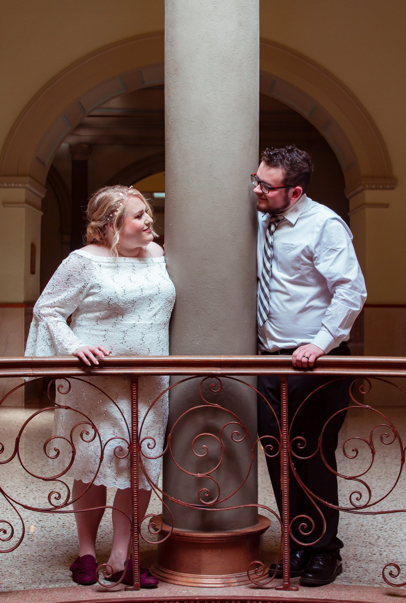 A man and a woman stand on either side of a large column, smiling at each other and leaning towards the column in a formal setting.