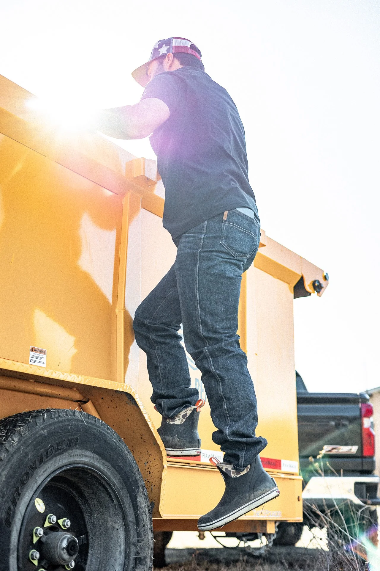 A person standing on the steps of a yellow utility truck, wearing jeans, a black shirt, and patterned high-top sneakers, with the sun shining brightly behind them.