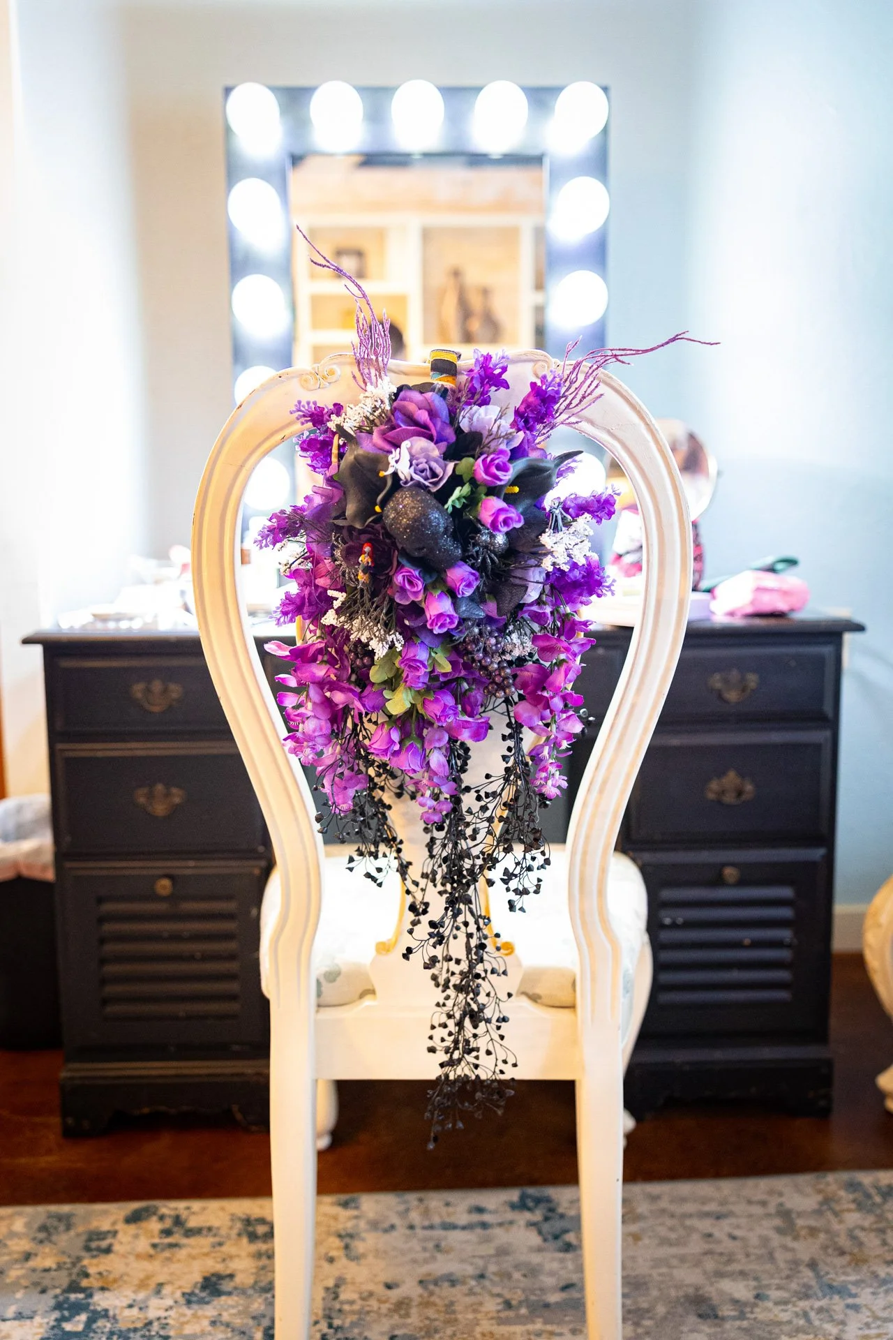 Elegant chair with a purple floral arrangement on the backrest, positioned in front of a mirror with bright round lights and a black dresser in a room.