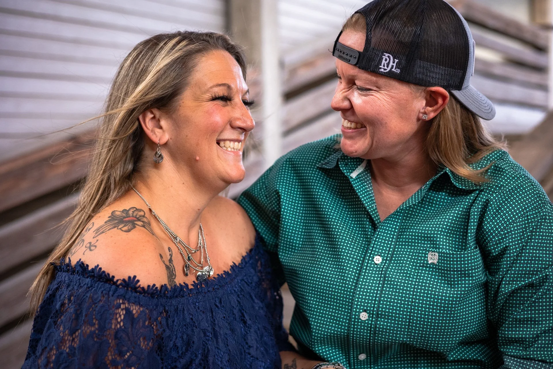 Two women smiling and laughing together, one wearing a blue off-shoulder top with tattoos and jewelry, the other wearing a green checkered shirt and a backwards baseball cap, sitting outdoors.