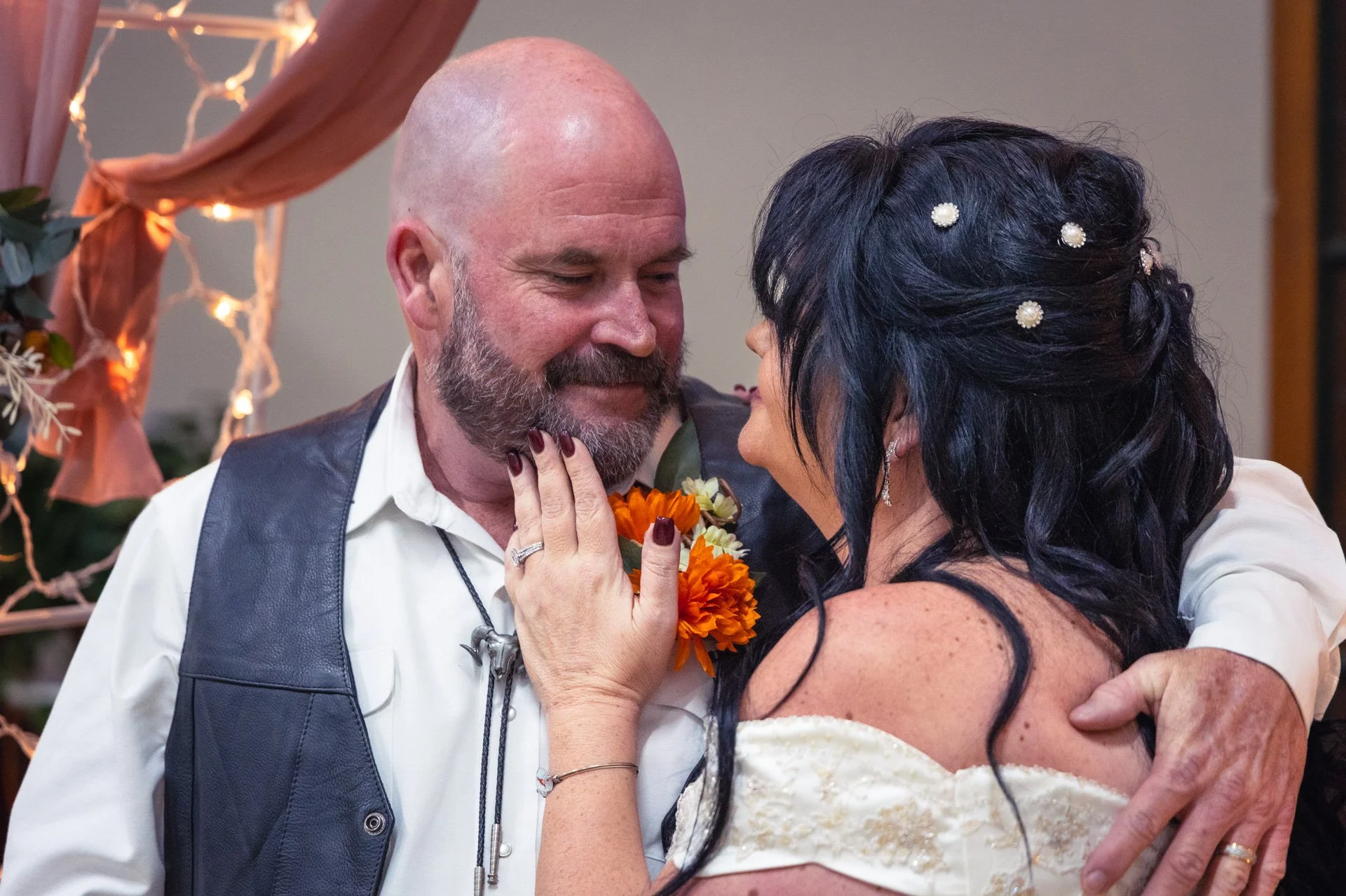 A couple sharing an intimate moment during their wedding, looking into each other's eyes with affectionate smiles. The man is bald with a beard, wearing a white shirt and a dark vest. The woman has long black hair adorned with small white decorative