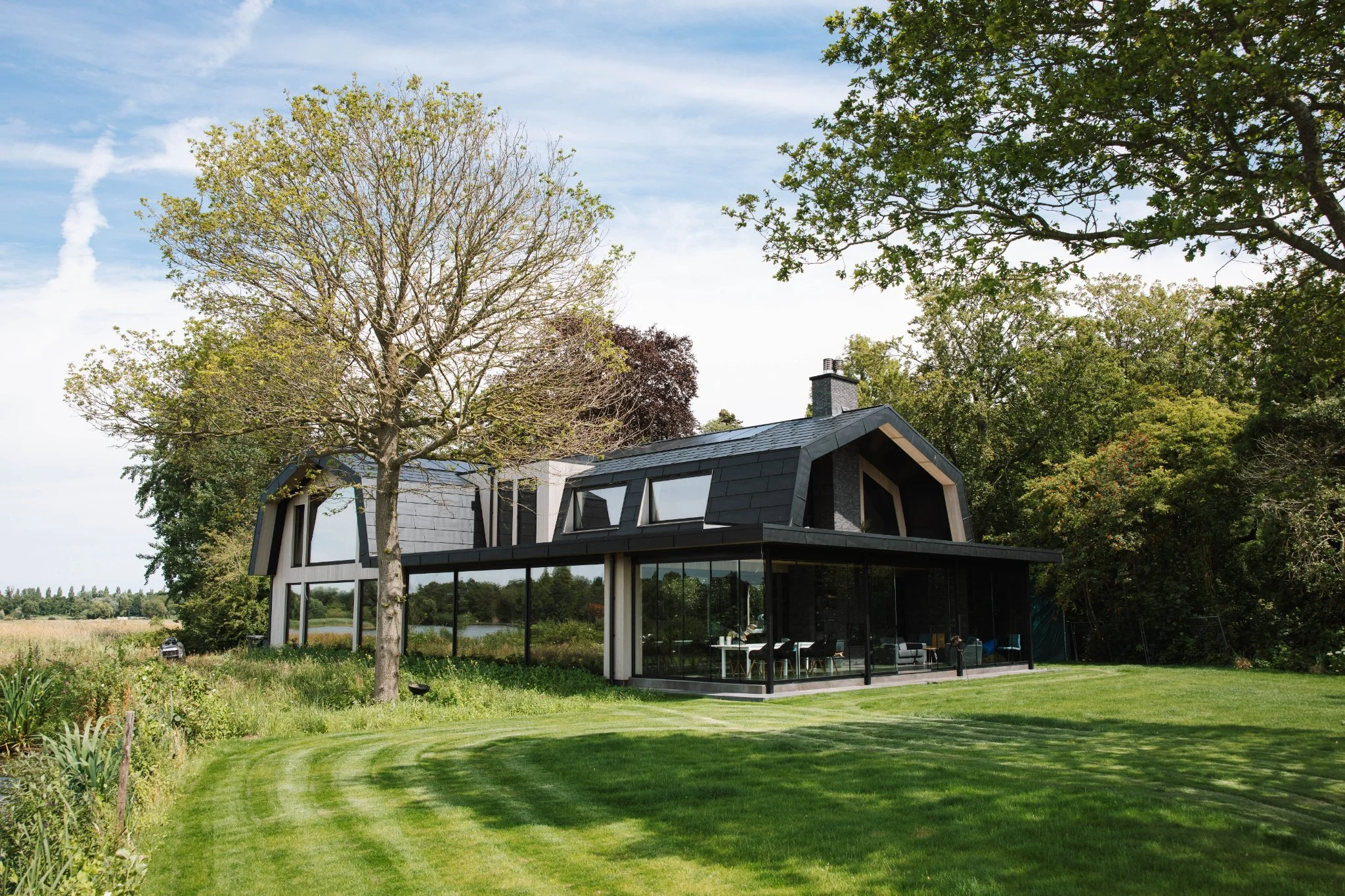 Modern huis met glazen wanden in een groene omgeving, omringd door bomen en een gazon, onder een blauwe hemel met enkele wolken.