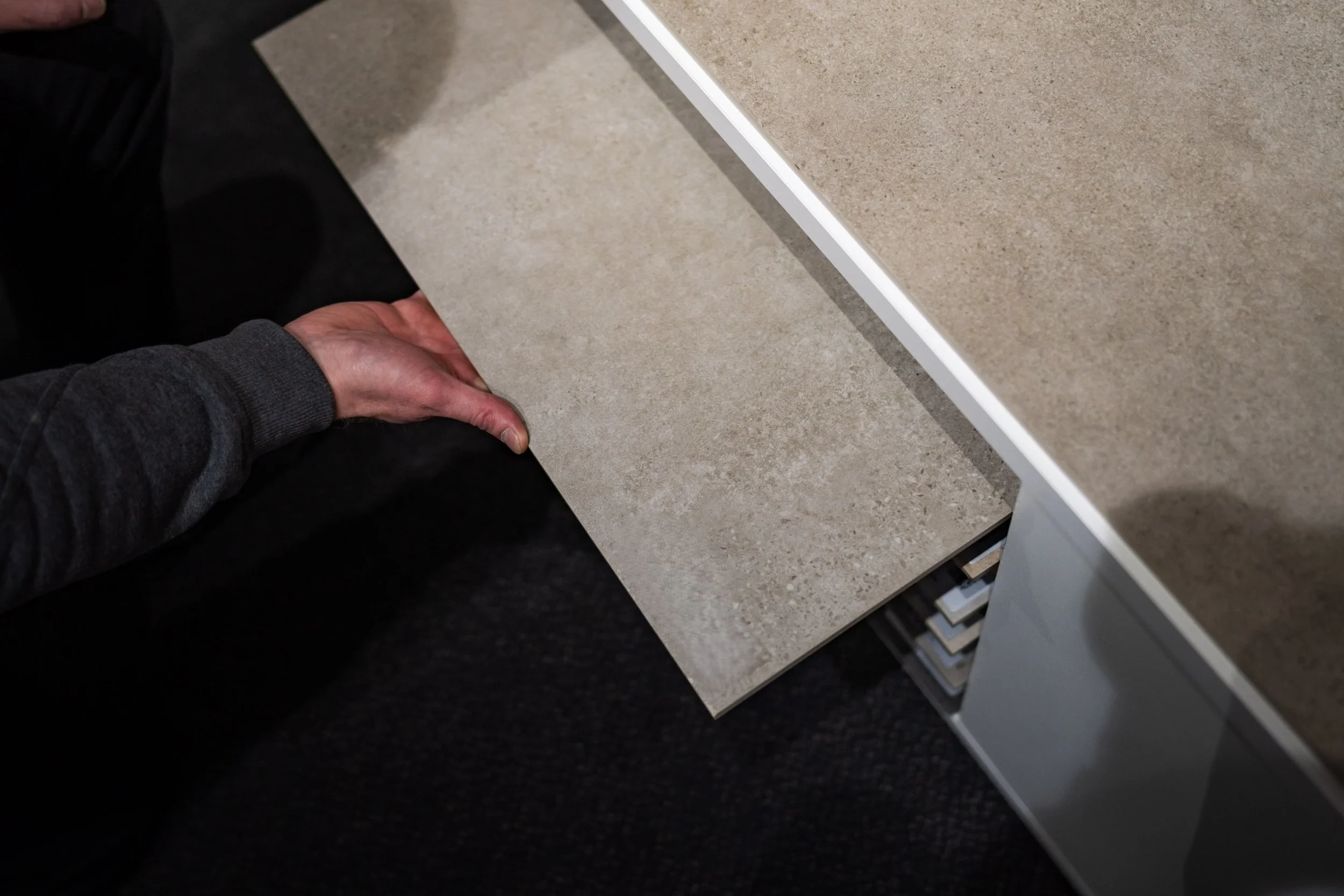 Richard Ensing houdt een tegel van beige keramiek vast naast een kast met tegelmonsters.