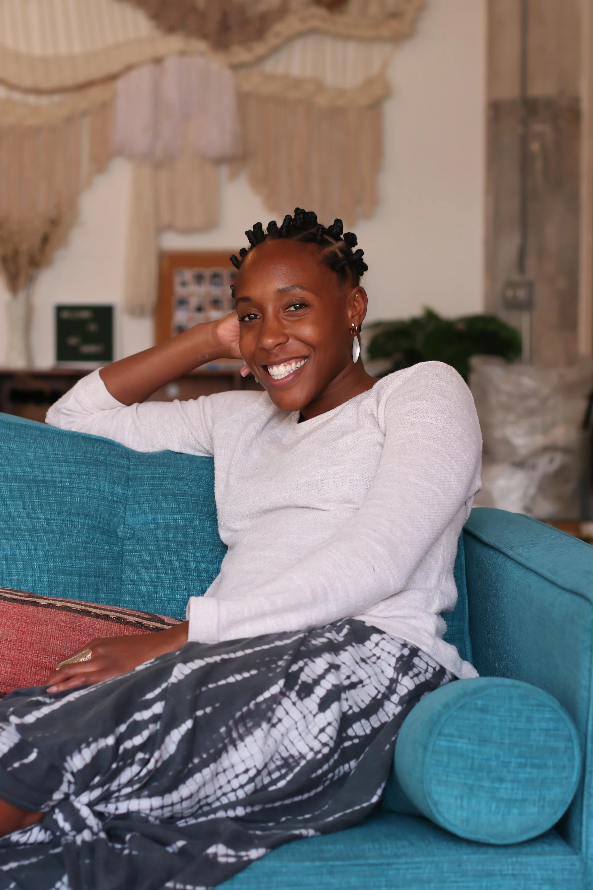 macabre wall background black woman sitting on teal couch beige sweater blue gray tie dye skirt