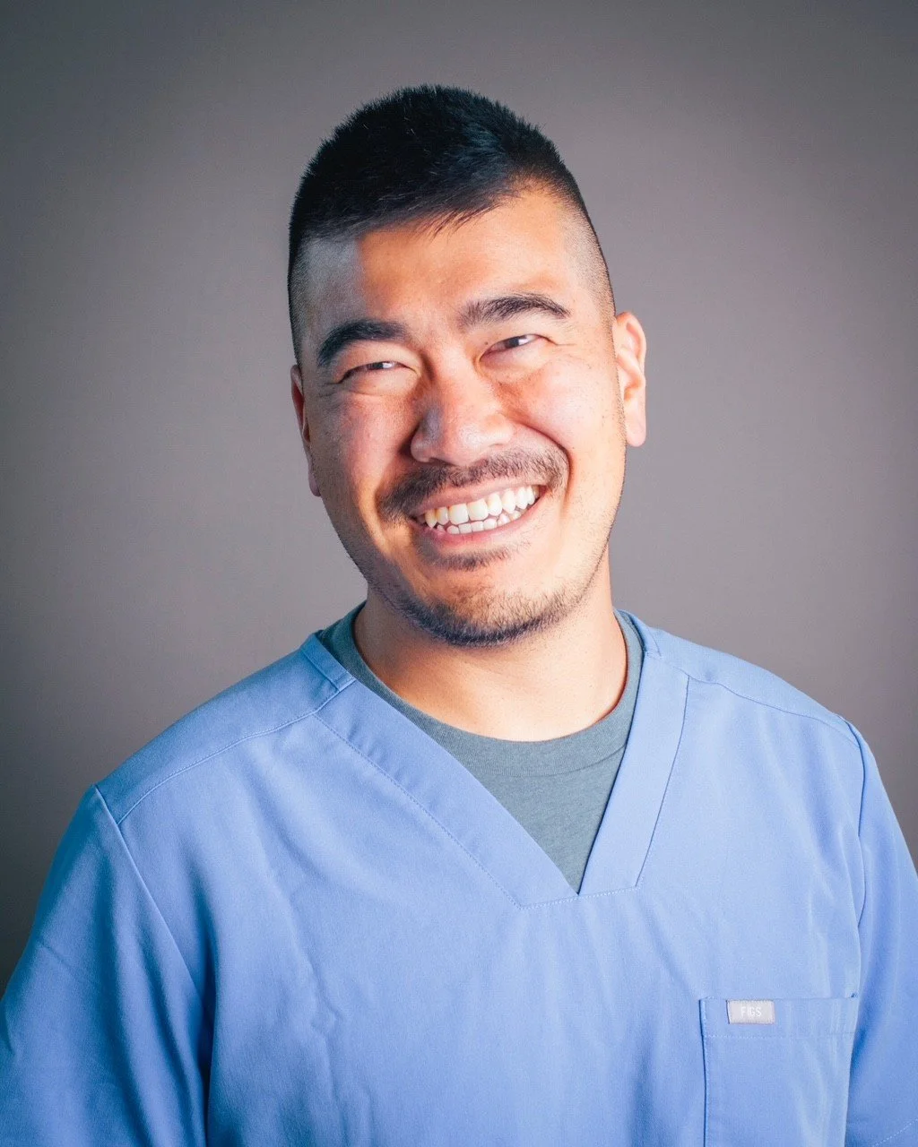 A man in light blue medical scrubs smiling with a gray background.