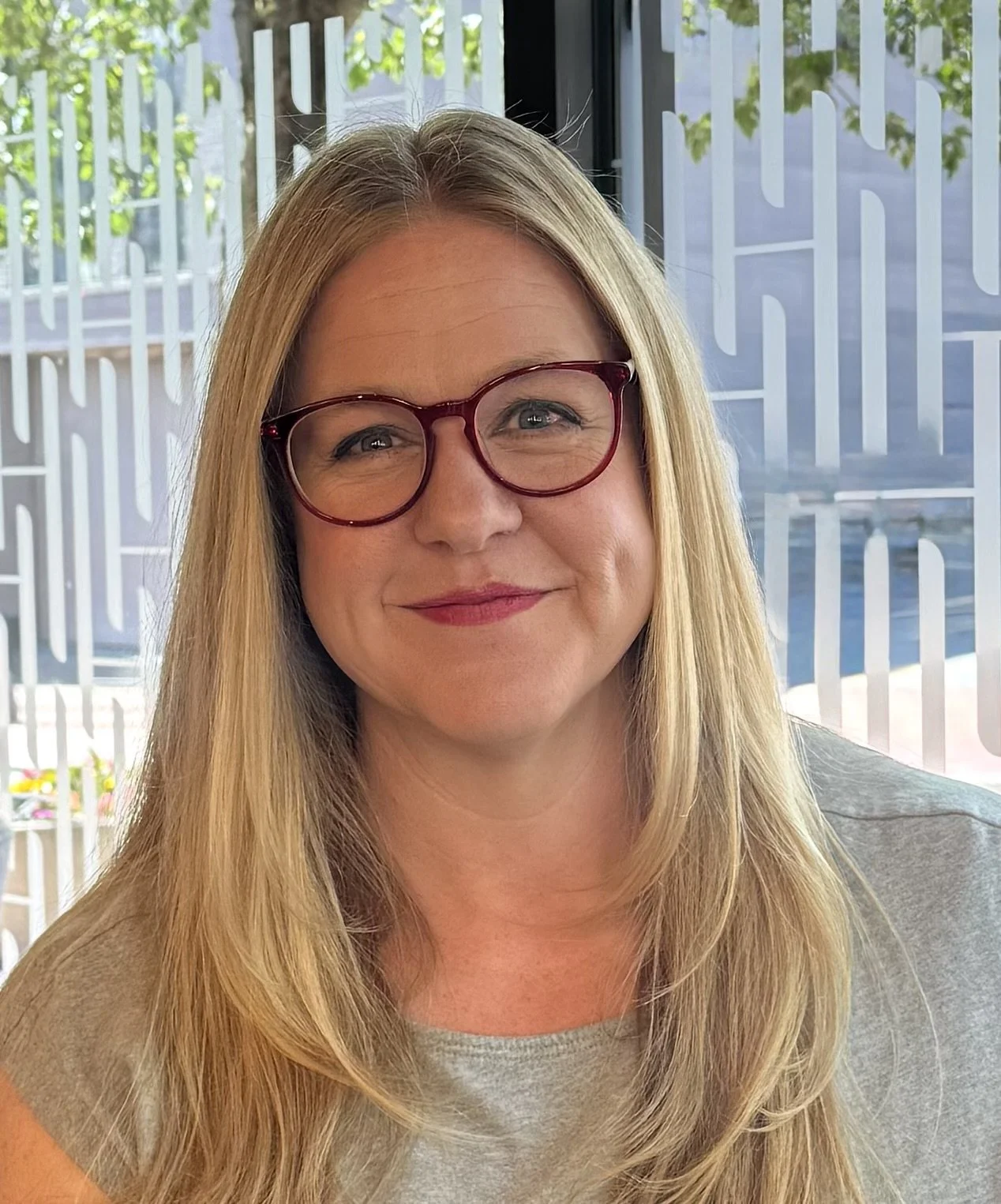 A woman with long blonde hair, wearing red glasses, smiling, in front of a window with patterned frosted glass, outside visible through the window