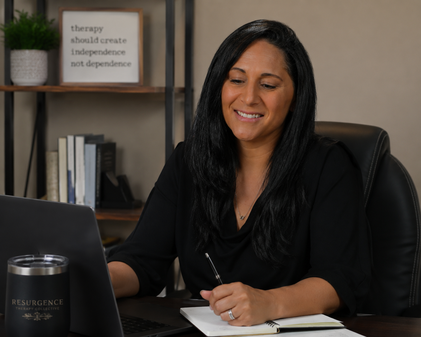 A woman with long black hair sitting at a desk, smiling, with a laptop, notebook, and pen. In the background, a bookshelf with books and a framed quote about therapy and independence.