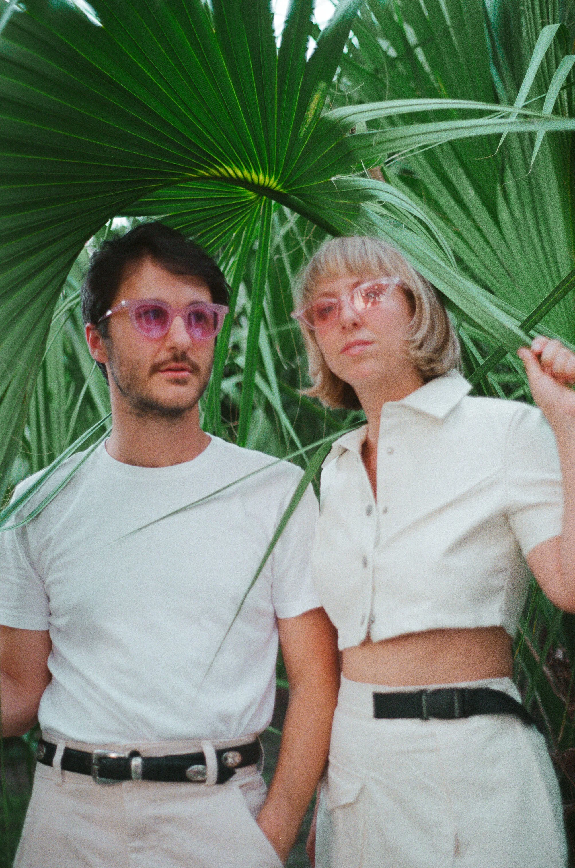 A young man and woman posing among large green tropical leaves, both wearing pink sunglasses and white clothing.