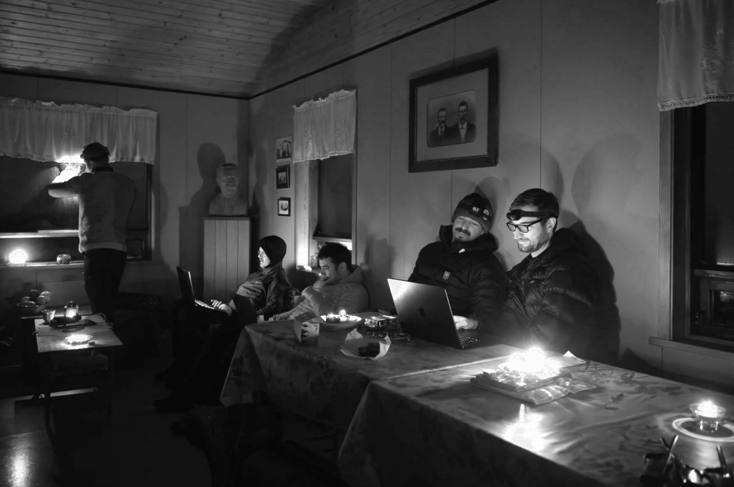 A black-and-white photo of six people in a cozy, dimly lit room with wooden walls. Four of them are seated at a table with laptops, and two are standing, one by a window. The walls have framed pictures and portraits, and small candles or lights illuminate the space softly.