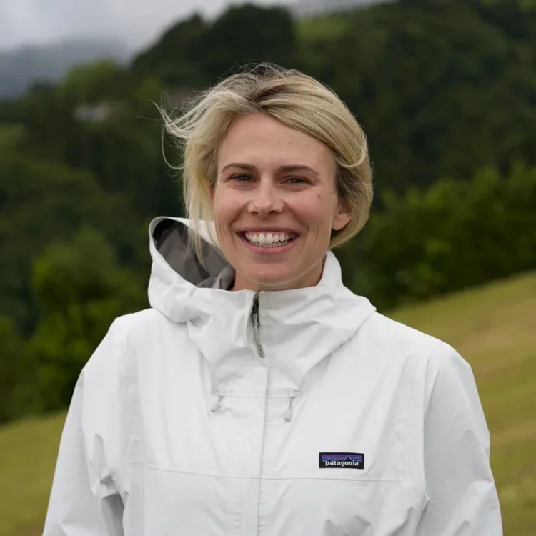 A woman smiling outdoors in a white Patagonia jacket with a green, hilly landscape in the background.