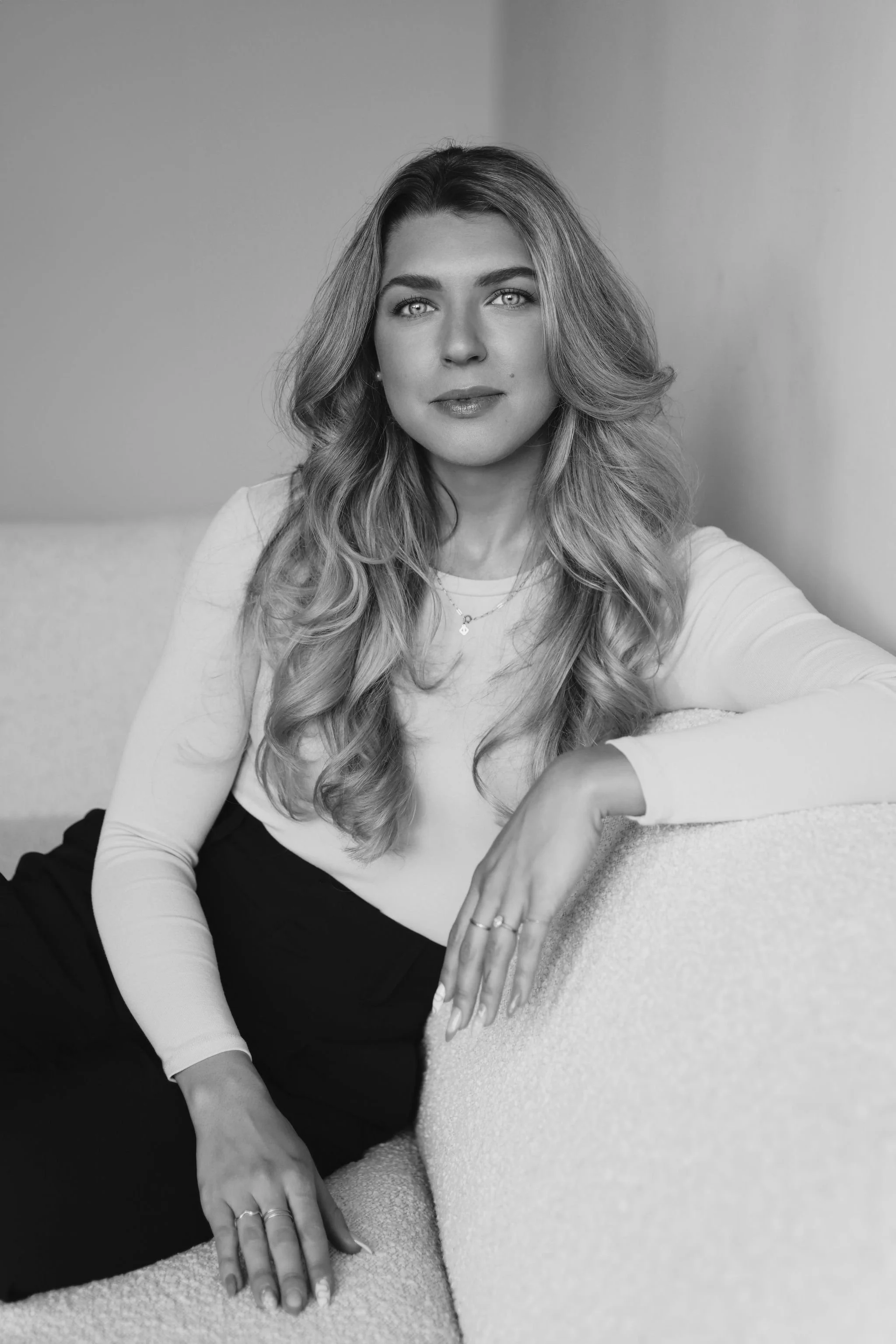 A woman with wavy blonde hair and light-colored eyes sitting on a sofa, looking at the camera in black and white