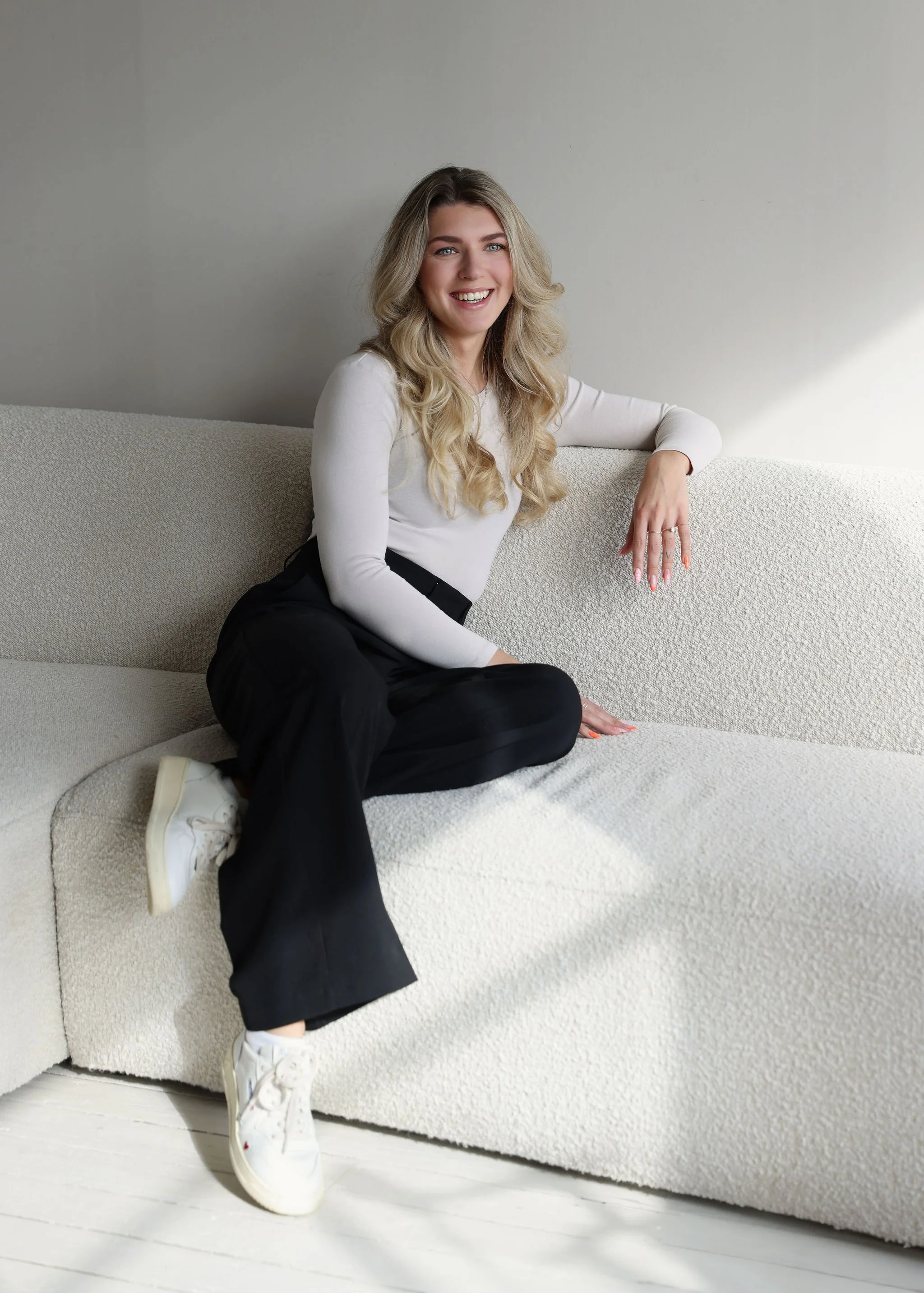 A smiling woman with long blonde hair sitting casually on a light-colored textured sofa, wearing a white long-sleeve top, black pants, and white sneakers. She is leaning her right arm on the back of the sofa and looking at the camera.